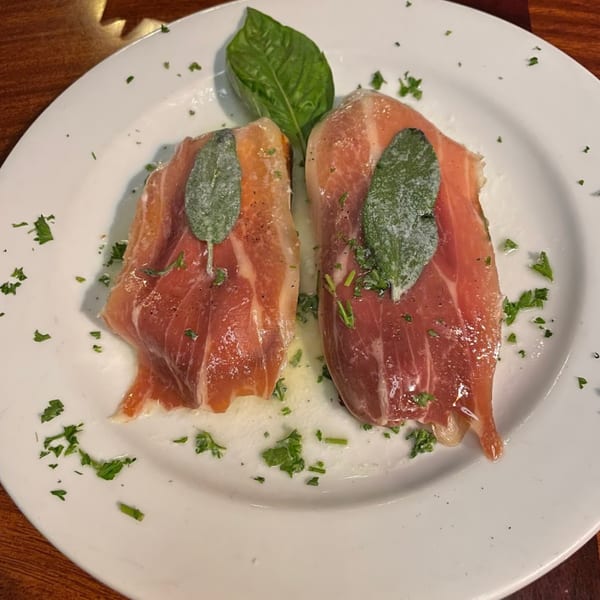 Two slices of prosciutto with sage leaves and parsley on a white plate.