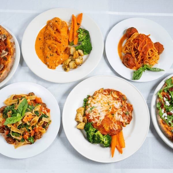 Overhead view of five Italian dishes, including pasta, pizza, and chicken entrees, on a white tablecloth.