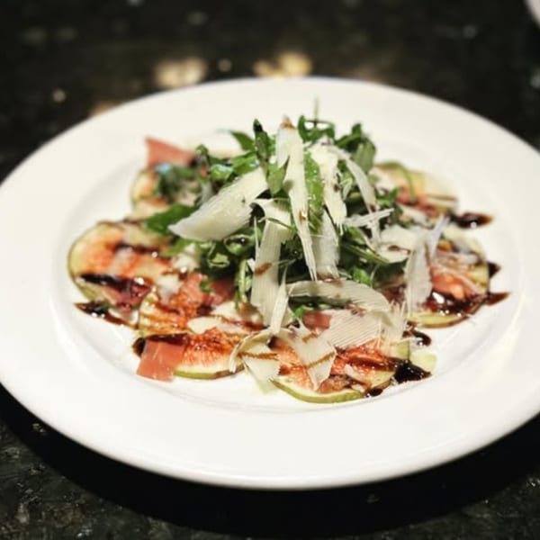 Italian fig and prosciutto appetizer with arugula, shaved parmesan, and balsamic glaze on a white plate.